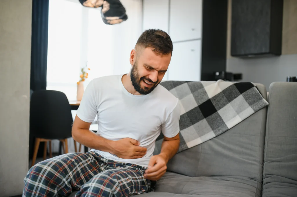 A man clutching his stomach in pain while sitting on a couch, showing symptoms of peptic ulcer disease (PUD).