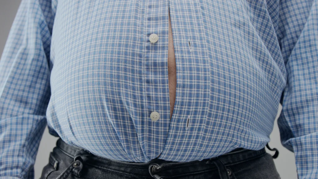 Close-up of a person wearing a blue checkered shirt with buttons straining due to bloating.