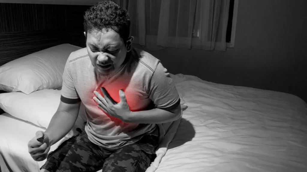 Man clutching chest in pain while sitting on the bed, showing nighttime discomfort from severe acid reflux or GERD symptoms.