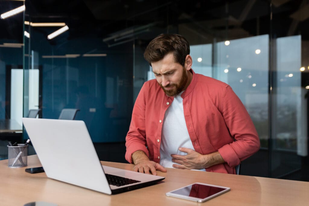 A man sitting at an office desk holding his stomach in pain, showing signs of digestive discomfort caused by stress.
