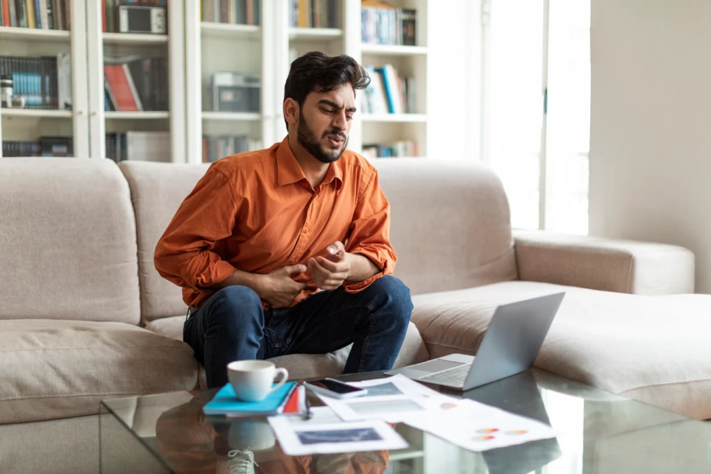 Man sitting on a couch holding his stomach in pain, possibly experiencing a Crohn’s Disease flare-up